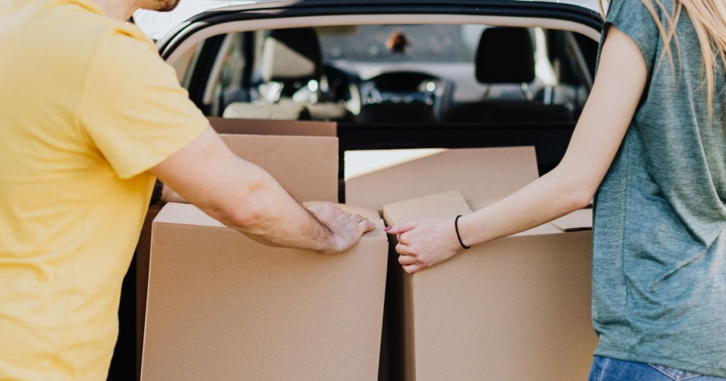 Two people loading large plain cardboard boxes into the open rear hatch of a personal passenger car parked outdoors on a sunny day.