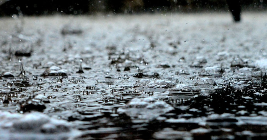 Heavy rain falling into standing water that has pooled and risen across the ground, with grass and trees blurred in the background.