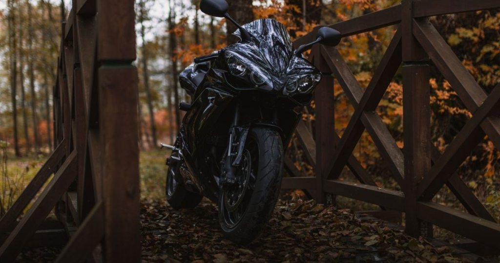 A black sport motorcycle parked on a wooden footbridge in the woods, surrounded by orange and brown fall leaves at the end of riding season.