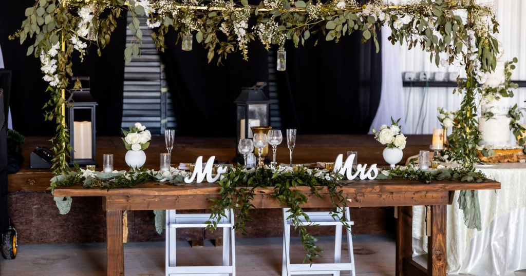 A rustic wooden sweetheart table with Mr and Mrs letters, greenery garland, white roses, lanterns and empty wine glasses set up inside a barn-style wedding venue, with a white wedding cake on a table to the right.