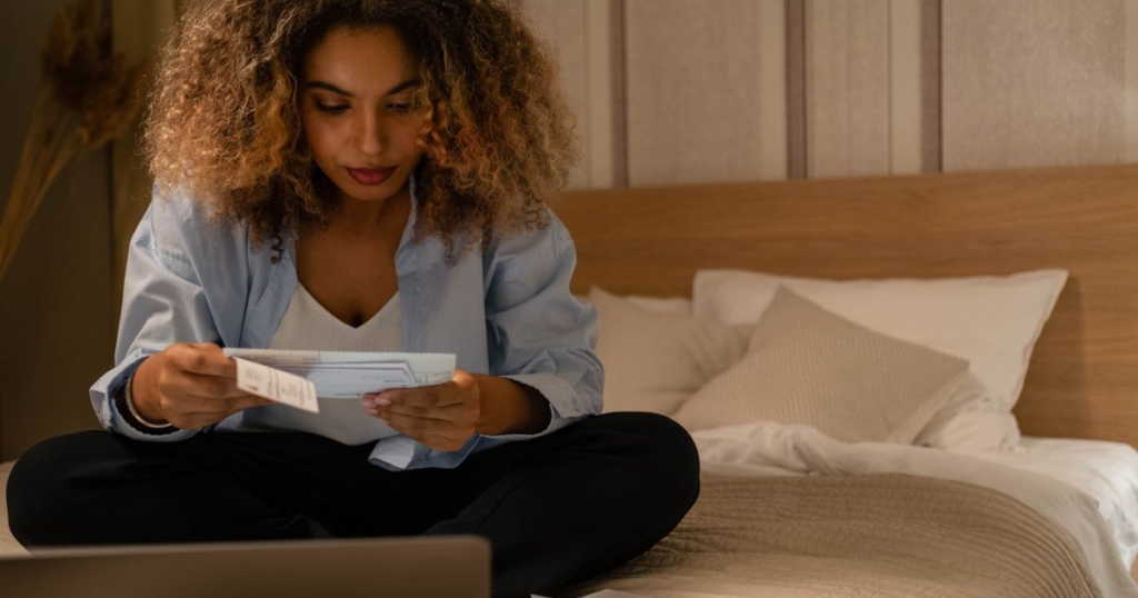 A woman sitting cross-legged on a bed at home, reading a printed notice she has just opened from the mail, with a laptop open in front of her.