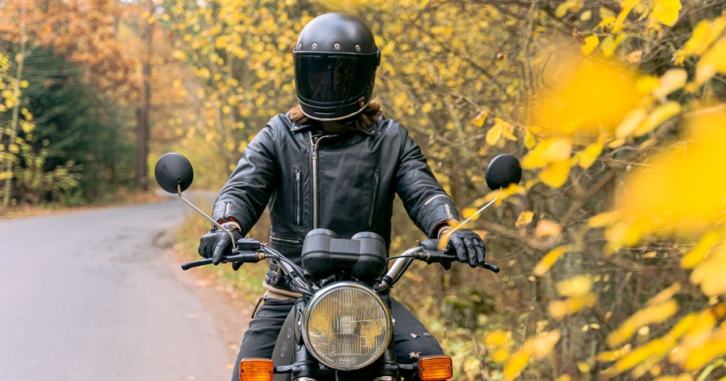Motorcycle rider in a helmet, leather jacket and gloves riding a two-lane road lined with yellow and orange autumn leaves
