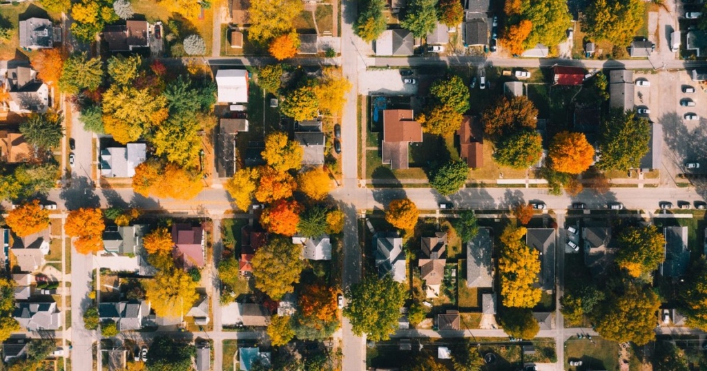 Overhead aerial view of an American residential neighborhood in autumn, with rows of single-family houses, driveways and parked cars along a grid of streets lined with orange and yellow trees.