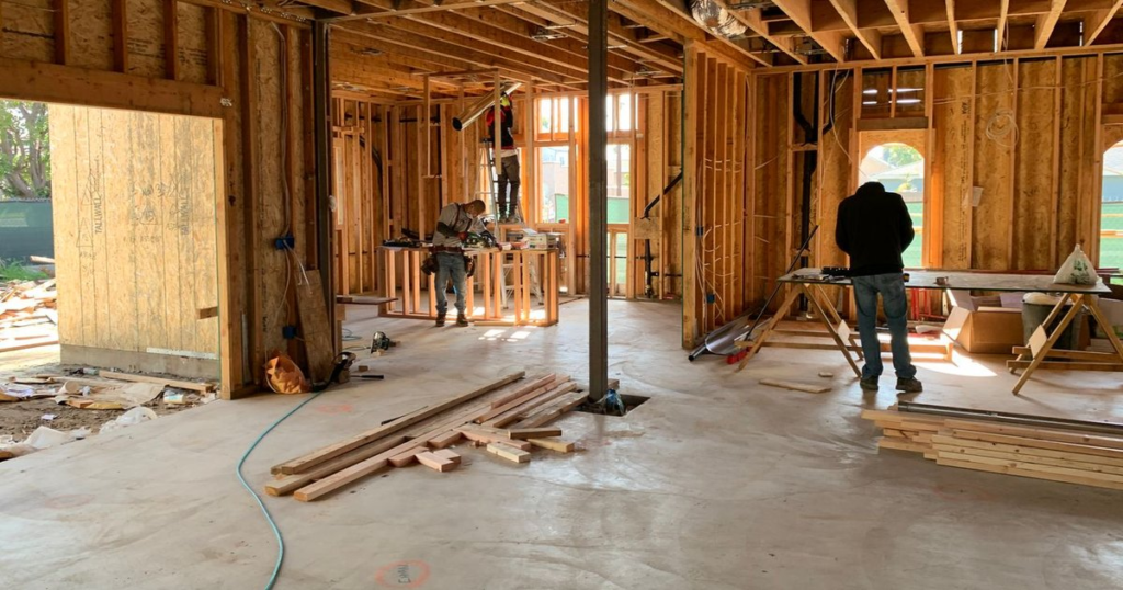 Three construction workers on the unfinished interior of a wood-framed house, one on a ladder overhead, one cutting lumber on a work table and one at a sawhorse bench.