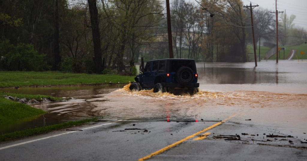 A dark blue Jeep Wrangler drives through muddy brown floodwater covering a two-lane road on a rainy, overcast day, with the yellow center line disappearing under the water and utility poles standing in the flooded field beyond.