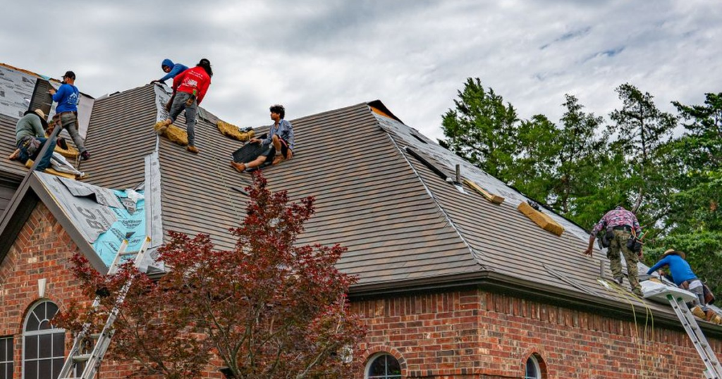 A six-person roofing crew works on the steep shingled roof of a red brick two-story house on an overcast day, with extension ladders leaning against the eaves and rolls of underlayment on the slope.