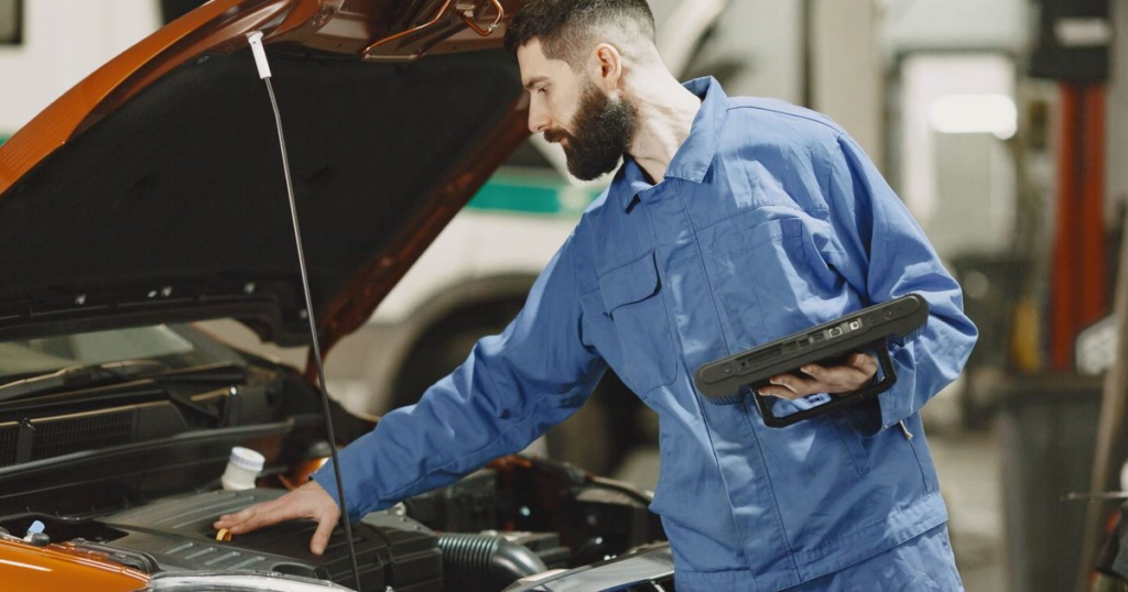 A technician in blue coveralls holding a handheld diagnostic tablet leans over the open hood of an orange car inside a repair shop.